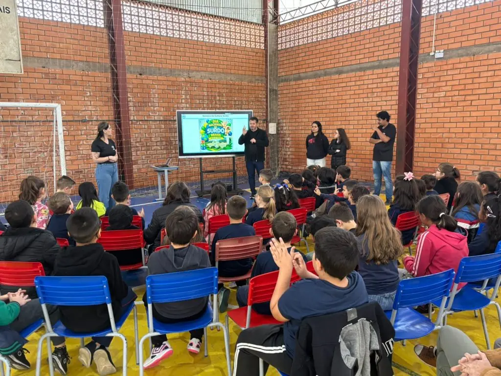 Equipe da APAS de Joaçaba apresenta atividade de Libras para alunos sentados em cadeiras no ginásio da Escola Municipal Nossa Senhora de Lourdes