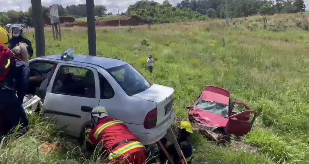 Bombeiros trabalham no desencarceramento de vítima em carro prata fora da pista, ao lado de outro veículo destruído