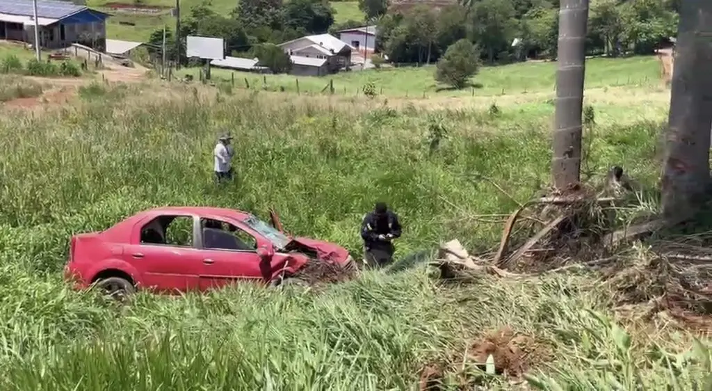Carro vermelho parado no gramado às margens da rodovia, com danos na frente