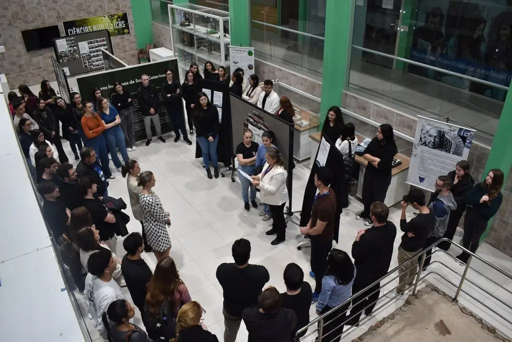 Vista do alto da abertura da exposição fotográfica, com público reunido entre os painéis no Museu do Centro Arqueológico da Unoesc Joaçaba