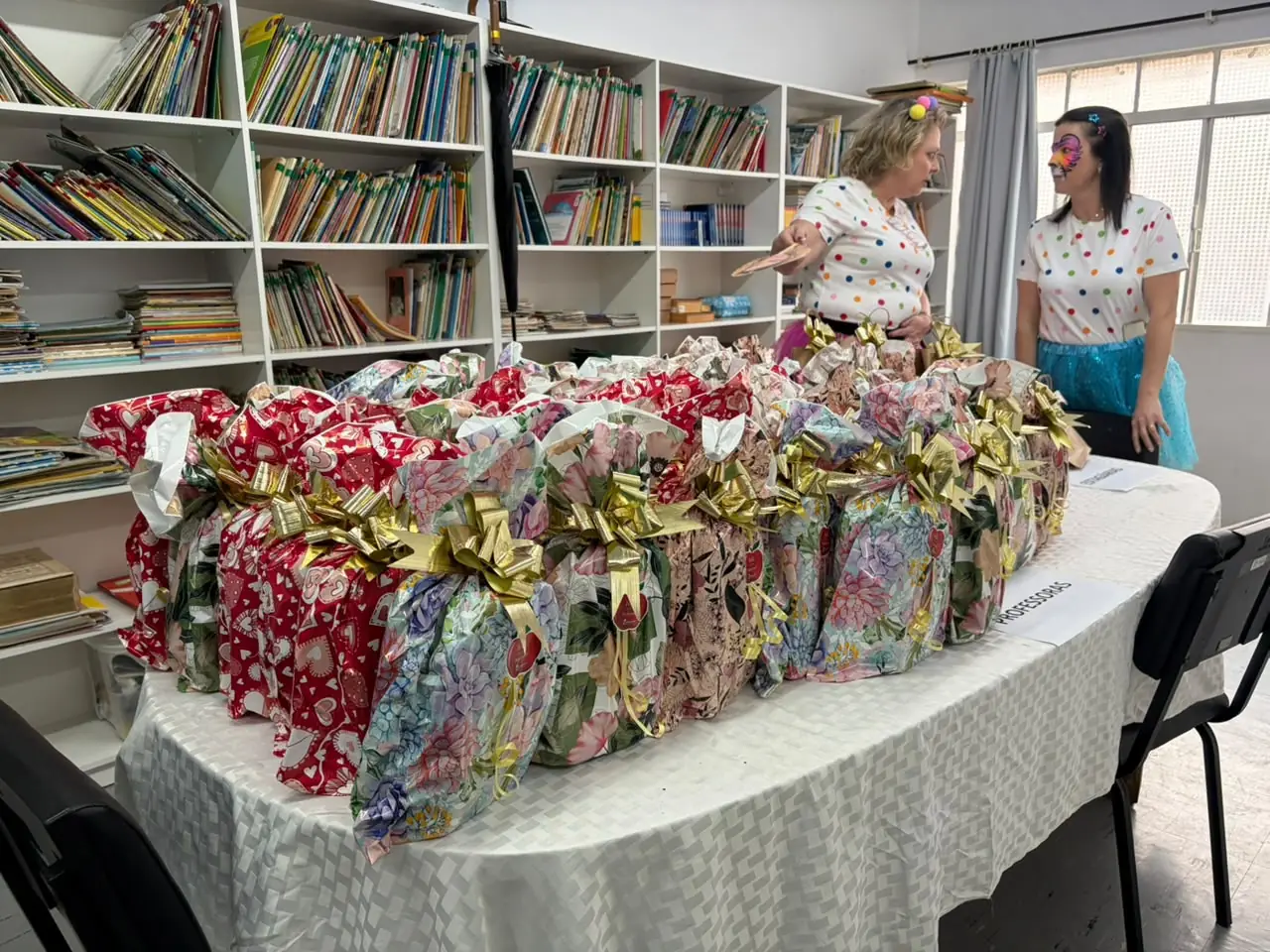 Presentes organizados sobre mesa na escola, com integrantes da CDL Mulher ao fundo