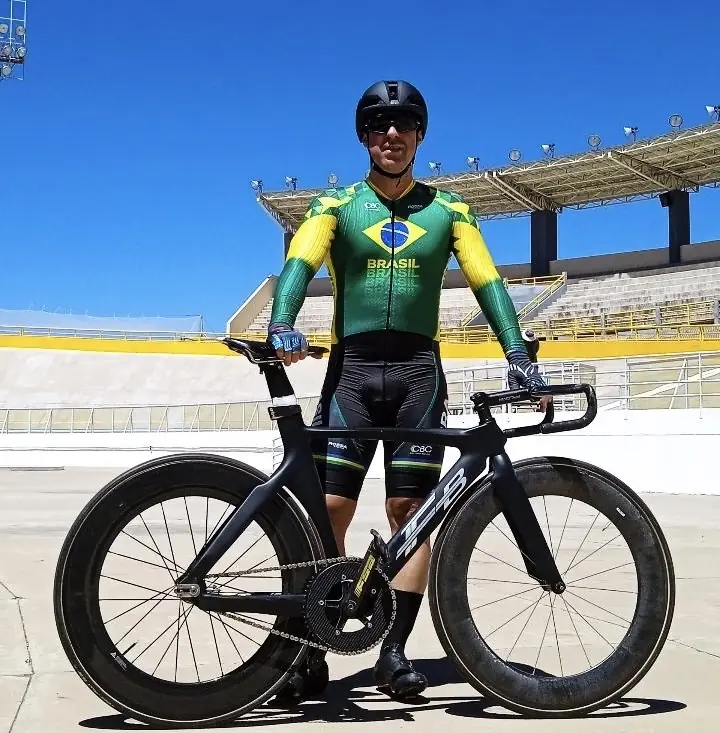 Ciclista com uniforme da Seleção Brasileira posa ao lado de bicicleta de pista, em velódromo
