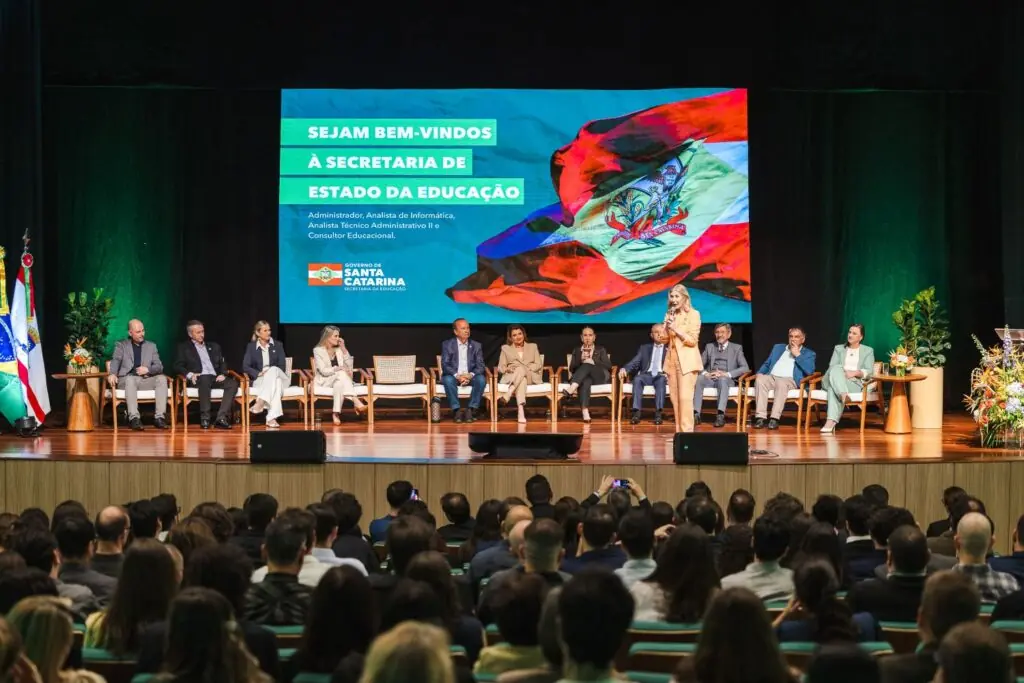 Secretária de Estado da Educação discursa no palco durante a cerimônia de posse dos novos servidores, diante do auditório lotado