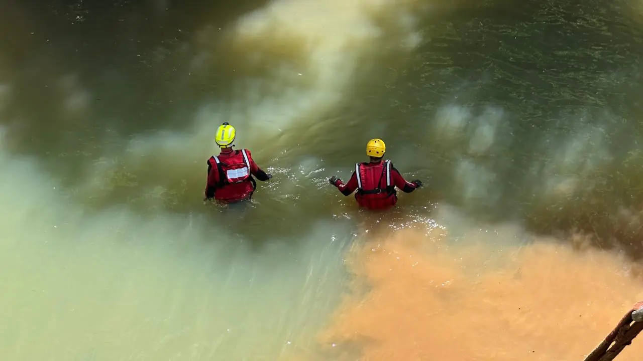 Dois bombeiros com coletes vermelhos e capacetes amarelos caminham dentro do Rio do Tigre