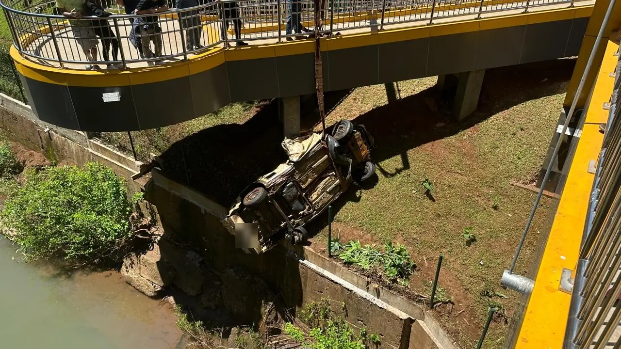 Carcaça do veículo suspensa por cinta ao lado da passarela, com pessoas observando