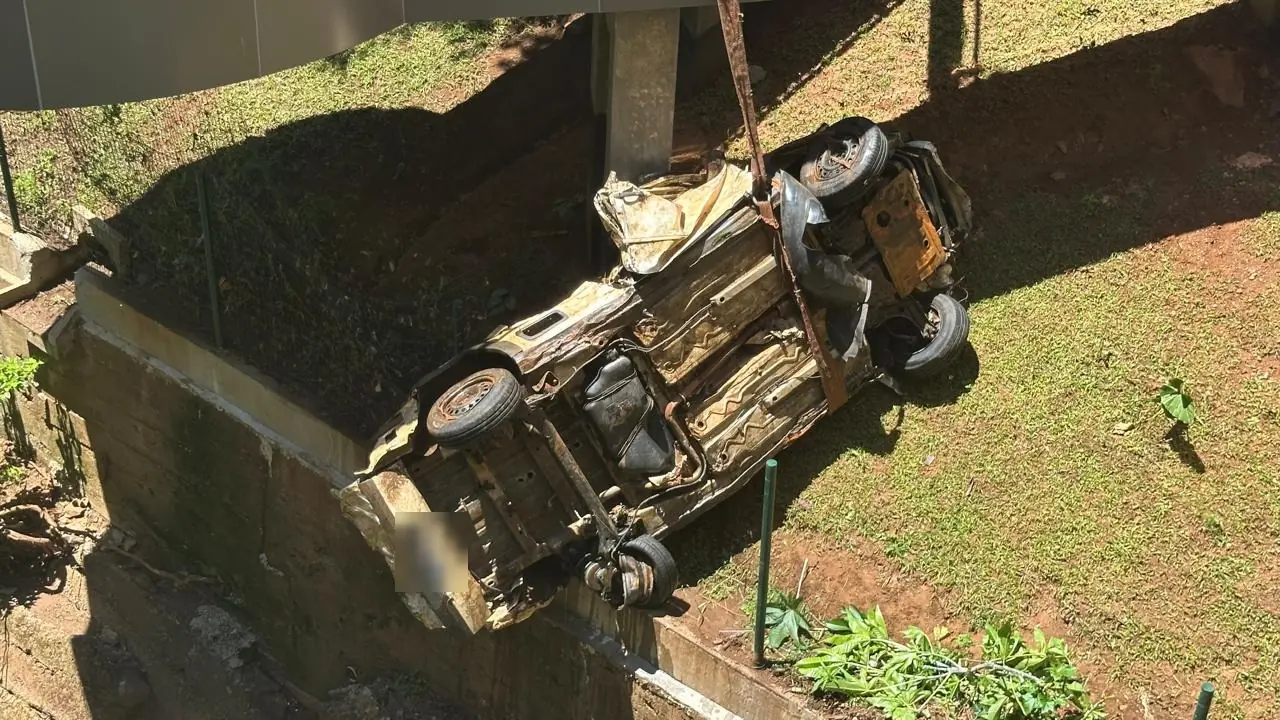 Carcaça do veículo virada de cabeça para baixo sobre o gramado, depois de ser retirada do rio